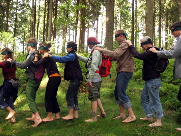 Mehrere Menschen gehen mit verbundenen Augen hintereinander. Sie haben ihre Hände auf die Schultern des Vorderen gelegt.