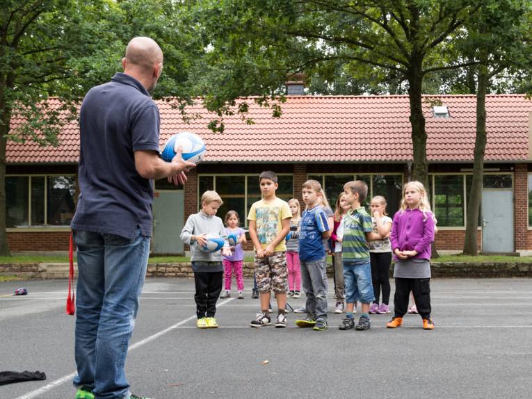 Ein Mann mit einem Rugby-Ei steht vor einer Gruppe mit Kindern.